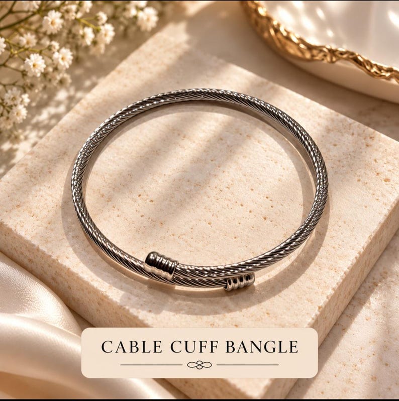 May include: A silver-toned cable cuff bangle featuring a twisted metal wire design. The bracelet has two smooth, cylindrical end caps that overlap slightly. The text CABLE CUFF BANGLE is printed in black on a rectangular label at the bottom.