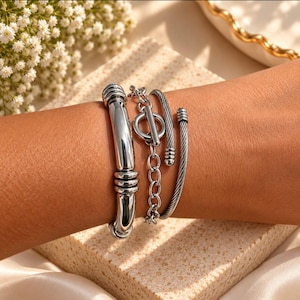 May include: A set of three silver-toned bracelets worn together. One is a smooth, thick polished bangle with ribbed accents, the second is a chain bracelet with a toggle clasp, and the third is a thin, twisted cable-style cuff with rounded ends.