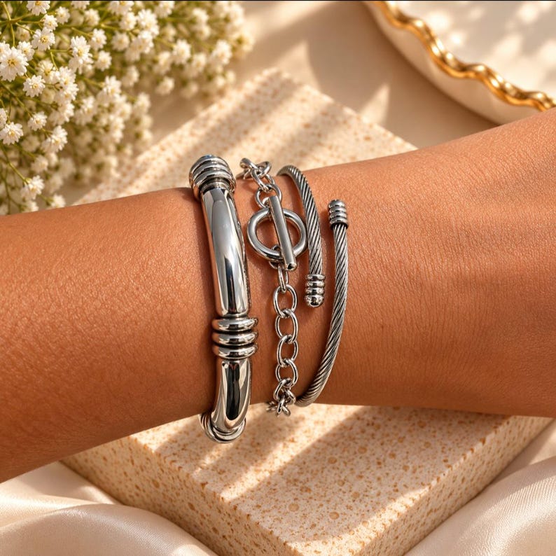 May include: A set of three silver-toned bracelets worn together. One is a smooth, thick polished bangle with ribbed accents, the second is a chain bracelet with a toggle clasp, and the third is a thin, twisted cable-style cuff with rounded ends.