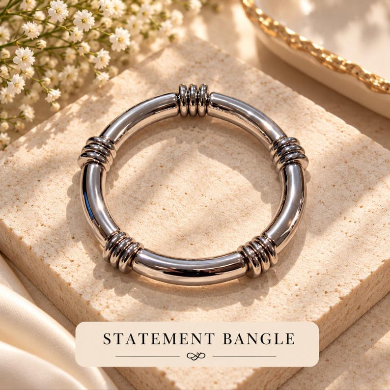 May include: A polished silver-tone metal bangle bracelet featuring a smooth, rounded band accented with four distinct sections of triple-ring detailing. The text "STATEMENT BANGLE" is printed in black on a rectangular label at the bottom.