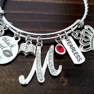 May include: Silver charm bracelet with multiple charms, including a heart, a book, a crown, a red gem, a tag that says "Fearless", and a tag that says "Black Girl Magic".