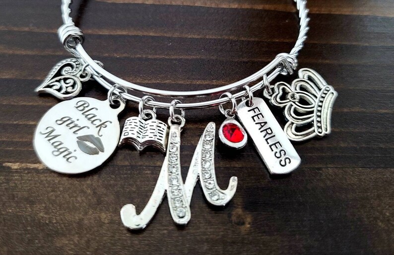 May include: Silver charm bracelet with multiple charms, including a heart, a book, a crown, a red gem, a tag that says "Fearless", and a tag that says "Black Girl Magic".