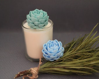 May include: A white candle in a glass jar with two blue succulent-shaped decorations on top. A bundle of dried green grass is in the foreground.