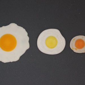 以下が含まれることがあります： 黒い表面に、大きさの異なる目玉焼きが3つ並んでいます。一番大きい目玉焼きはオレンジ色の黄身、真ん中の目玉焼きは黄色の黄身、一番小さい目玉焼きはオレンジ色の黄身です。