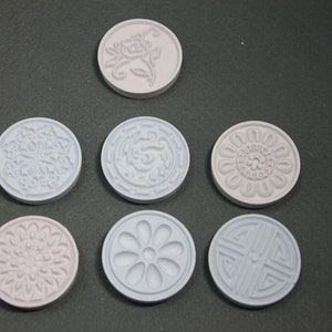 May include: Seven round, decorative, ceramic stamps with raised designs. The stamps are in shades of light purple and light blue. The designs include a flower, a geometric pattern, and a stylized letter.