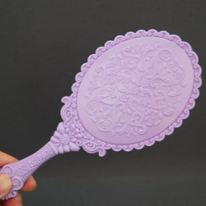 May include: A purple hand mirror with a decorative floral design. The mirror has a scalloped edge and a handle with a floral design.