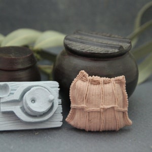 May include: A set of three miniature pottery items: a small brown pot with a lid, a light grey miniature stove with a pot on top, and a light brown fabric sack with a woven pattern.