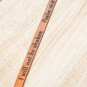 May include: A brown leather bracelet with the words "I will not be shaken" and "Psalm 16:8" engraved on it.