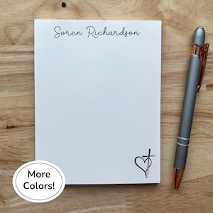 Peut inclure: Un bloc-notes blanc avec le nom "Soren Richardson" en écriture cursive, un motif cœur et croix, et un stylo gris avec des accents cuivrés. Le bloc-notes est sur une surface en bois, avec un bouton "More Colors!" en bas à gauche.