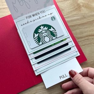 Pu&ograve; includere: Un porta carte rosso con un inserto bianco che dice "FOR WHEN YOU need a pick me up". Il porta carte contiene una carta regalo Starbucks e altre carte. La parola "PULL" &egrave; visibile sull'inserto inferiore.