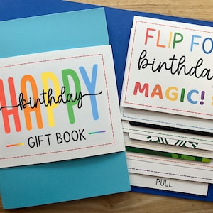 Pu&ograve; includere: Un colorato libro di regali di compleanno con una copertina blu. La copertina recita "HAPPY birthday GIFT BOOK" in colori arcobaleno. All'interno, le carte dicono "FLIP FOR birthday MAGIC!" e "PULL".