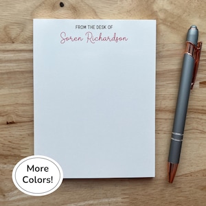 Peut inclure: Un bloc-notes blanc avec le texte "FROM THE DESK OF Soren Richardson" en script rouge. Un stylo gris avec des accents or rose repose sur une surface en bois. Le bloc-notes est vierge, prêt pour les notes.
