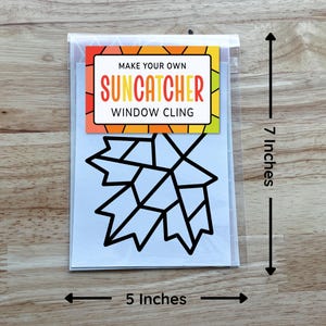 Op de afbeelding: Een Make Your Own Suncatcher raamsticker-set met een zwarte omtrek van een esdoornblad op een witte achtergrond. Het verpakkingslabel heeft kleurrijke geometrische randen en meet 17,78 cm hoog bij 12,7 cm breed.