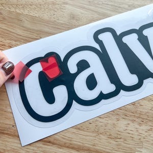 K&ouml;nnte beinhalten: Ein wei&szlig;er Vinyl-Aufkleber mit dem Wort "Calvin" in schwarzen Buchstaben. Der Aufkleber wird von einem wei&szlig;en Blatt Papier abgezogen.