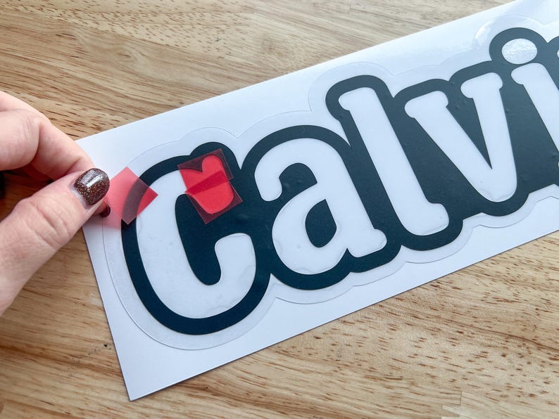 K&ouml;nnte beinhalten: Ein wei&szlig;er Vinyl-Aufkleber mit dem Wort "Calvin" in schwarzen Buchstaben. Der Aufkleber wird von einem wei&szlig;en Blatt Papier abgezogen.
