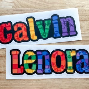 K&ouml;nnte beinhalten: Zwei regenbogenfarbene Vinyl-Aufkleber mit den Namen "Calvin" und "Lenora" in schwarz umrandeten Buchstaben.