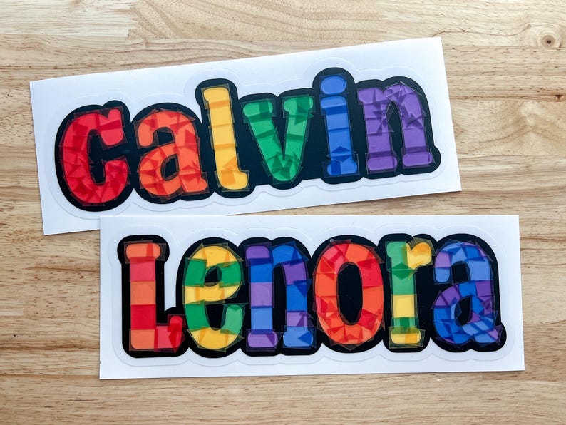 K&ouml;nnte beinhalten: Zwei regenbogenfarbene Vinyl-Aufkleber mit den Namen "Calvin" und "Lenora" in schwarz umrandeten Buchstaben.