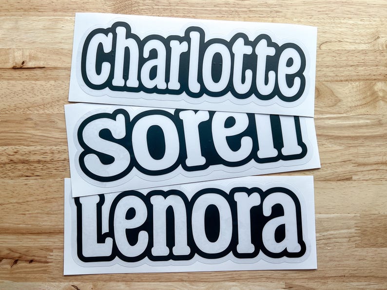 K&ouml;nnte beinhalten: Drei wei&szlig;e Aufkleber mit schwarzem Text. Der obere Aufkleber sagt "charlotte", der mittlere Aufkleber sagt "sorel" und der untere Aufkleber sagt "lenora".