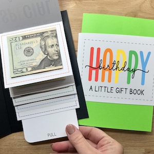 May include: A birthday gift book with a twenty-dollar bill visible. The cover reads "HAPPY birthday A LITTLE GIFT BOOK" in colorful letters. The book has a pull-out section with the word "PULL" on it.