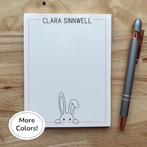 May include: A white notepad with a pink border and the name "CLARA SINNWELL" at the top. A black and white bunny illustration is at the bottom. A gray pen with rose gold accents rests on a wooden surface.