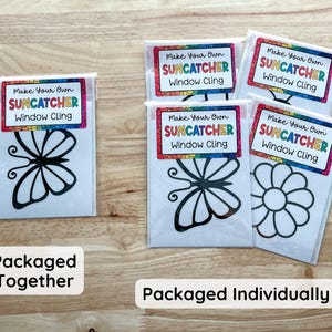 May include: Clear plastic packages containing suncatcher window clings with various designs, including a butterfly and a flower. Each package has a label with the text "Make Your Own Suncatcher Window Cling". The packages are on a wooden surface.