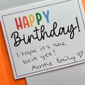 Pu&ograve; includere: Un biglietto di compleanno con le parole "HAPPY Birthday!" in lettere colorate. Il biglietto include un messaggio scritto a mano che dice "I hope it's the best yet! Auntie Emily" con un simbolo di cuore. Il biglietto &egrave; all'interno di una busta arancione.