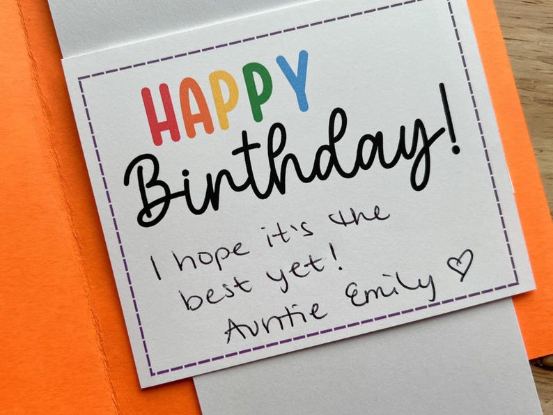 Pu&ograve; includere: Un biglietto di compleanno con le parole "HAPPY Birthday!" in lettere colorate. Il biglietto include un messaggio scritto a mano che dice "I hope it's the best yet! Auntie Emily" con un simbolo di cuore. Il biglietto &egrave; all'interno di una busta arancione.