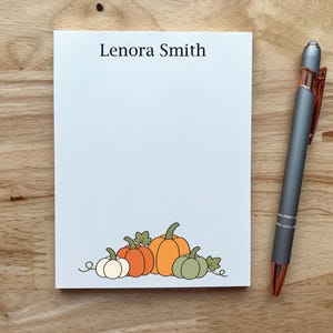 May include: A white rectangular notepad with the name Lenora Smith printed in black at the top and a graphic of four pumpkins in orange, white, and sage green at the bottom, accompanied by a grey and copper-colored retractable ballpoint pen.