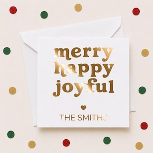 May include: A square white greeting card with a matching envelope featuring gold foil text that reads merry happy joyful and THE SMITHS with a small gold heart icon centered between the text and the family name on a light background.