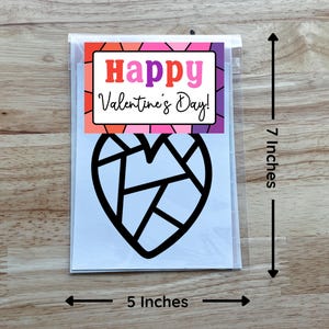May include: A Valentine's Day card in a clear plastic sleeve. The card features a black geometric heart design and the words "Happy Valentine's Day!" in a colorful frame. The card measures 5 inches wide and 7 inches tall.
