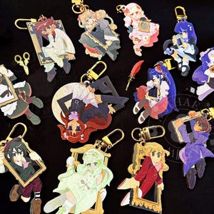 Puede incluir: Una colección de doce llaveros de acrílico con varios personajes de estilo anime, cada uno enmarcado en un borde decorativo. El conjunto incluye un pequeño dije de tijeras de color dorado y una pluma roja, unidos a cierres de mosquetón dorados.