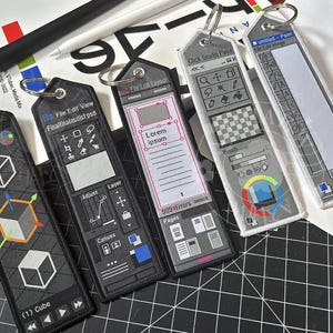 May include: Five black and white embroidered keyrings featuring software interface designs. The keyrings include designs for Blender, Photoshop, and other digital art programs. Each keyring has a silver ring for attachment.