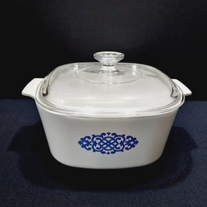 May include: White casserole dish with a clear glass lid and handles. The dish features a blue decorative pattern on the front. The lid has a small knob for easy handling. A classic piece for serving.