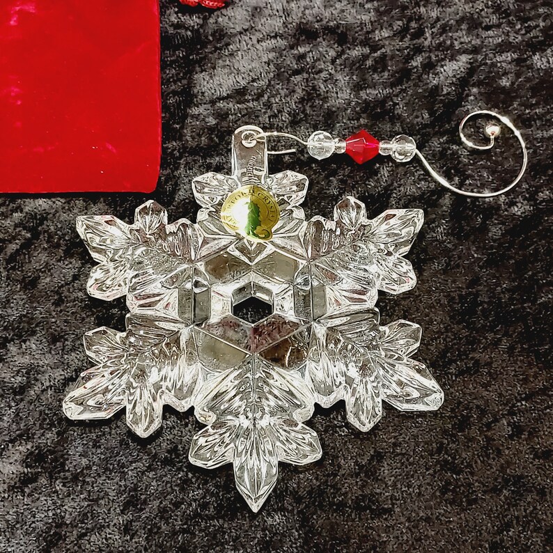 K&ouml;nnte beinhalten: Ein klarer Glas-Schneeflocken-Anh&auml;nger mit einem goldfarbenen Metall-H&auml;nger und einer roten Perle. Die Schneeflocke ist mit filigranen Kristallmustern verziert.