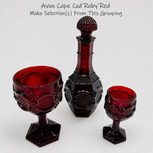 May include: Avon Cape Cod Ruby Red glassware set featuring a decanter with a textured stopper and two goblets of varying sizes. Each piece is crafted from deep red glass with a faceted, geometric pattern and a hexagonal base design.