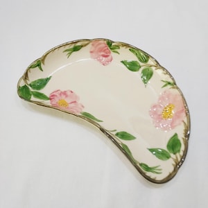 May include: Off-white, fan-shaped ceramic serving dish with pink floral and green leaf designs. The rim is trimmed with a gold-colored border. The dish has a scalloped edge and is likely used for serving appetizers or desserts.