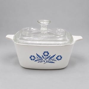 May include: A rectangular white casserole dish with integrated side handles and a clear glass lid. The front of the dish features a blue floral pattern consisting of two stylized flowers with pointed petals and central leaves.
