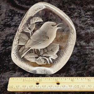 May include: A clear glass paperweight with a detailed etched design of a bird perched on a branch with leaves. The paperweight is shaped like a rounded oval.