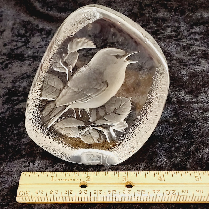 May include: A clear glass paperweight with a detailed etched design of a bird perched on a branch with leaves. The paperweight is shaped like a rounded oval.