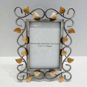 May include: A silver-toned metal picture frame with gold-colored leaf accents. The frame is designed with swirling lines and decorative leaves. The text on the frame reads "Sixtrees Gold Leaf & Silver Tone Frame 3-1/2" x 5" Image."
