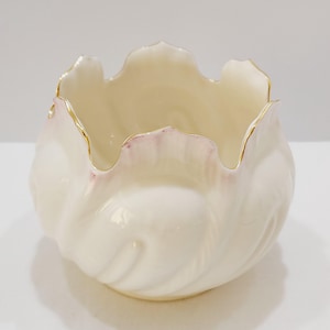 May include: A cream-colored, ceramic bowl with a scalloped rim. The rim is trimmed with gold and has a subtle pink hue. The bowl's interior is a soft cream color, and the exterior has a textured, petal-like design.