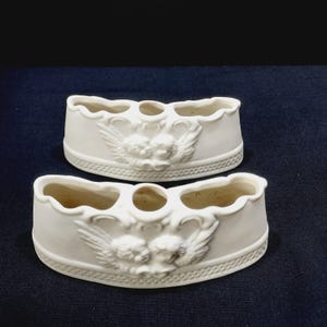 May include: Two white ceramic toothbrush holders. Each holder has a curved shape with three openings and a decorative relief of cherubs with wings. The holders have a scalloped edge and a textured base.