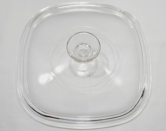 May include: A square, clear glass lid with rounded corners and a centered, circular knob handle. The lid features a stepped, recessed rim design intended to fit securely onto a matching square container or pan, providing a transparent view.