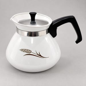 May include: A white enamel teapot featuring a brown wheat stalk graphic on the side. The teapot has a black handle, a silver-toned metal band around the neck, and a silver lid with a small black knob handle on top for easy lifting and pouring.