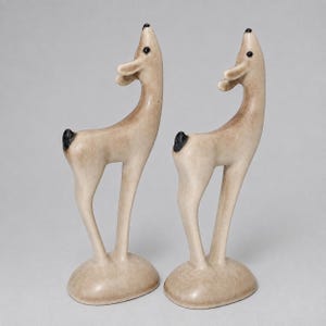 May include: A pair of standing deer figurines crafted from a smooth, light beige material. Each deer features a long, slender neck, a small black tail, and black painted eyes. The figures are positioned on oval bases, facing upward with ears extended.