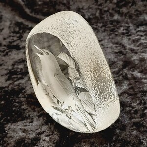 May include: A clear glass paperweight with a detailed etched design of a bird singing in a tree. The glass has a textured surface.