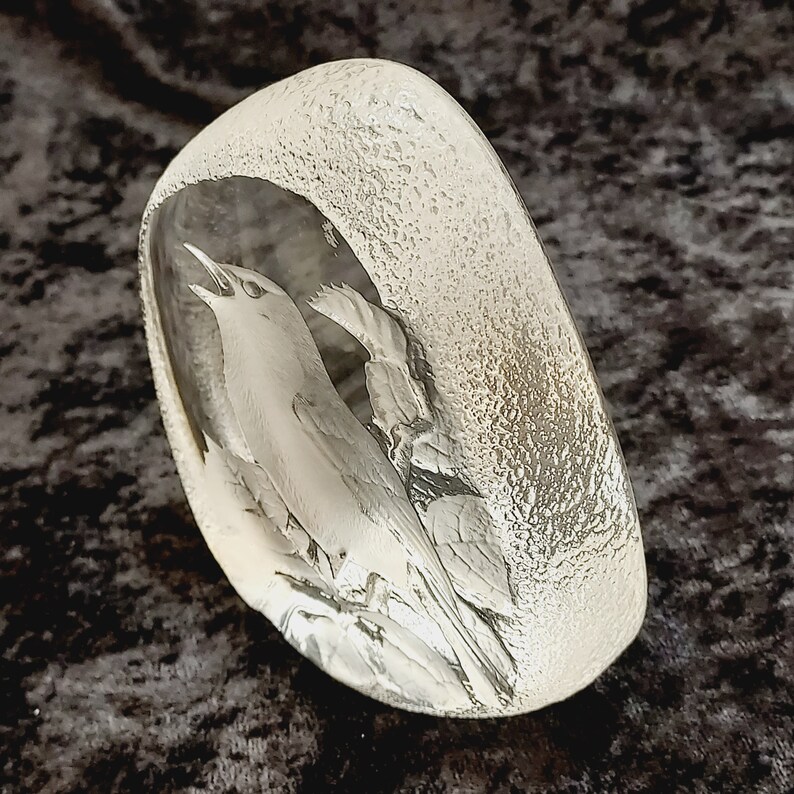 May include: A clear glass paperweight with a detailed etched design of a bird singing in a tree. The glass has a textured surface.