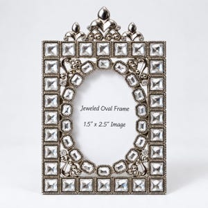 May include: A rectangular silver-toned picture frame featuring a central oval opening surrounded by clear square and rectangular crystals. The frame includes a decorative top crest and text reading Jeweled Oval Frame 1.5" x 2.5" Image.