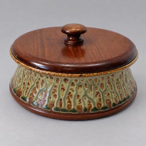 May include: A round ceramic container with a dark brown wooden lid featuring a central knob. The base is glazed in a textured, mottled pattern of sage green and earthy brown tones, creating a distinct organic appearance on the exterior surface.