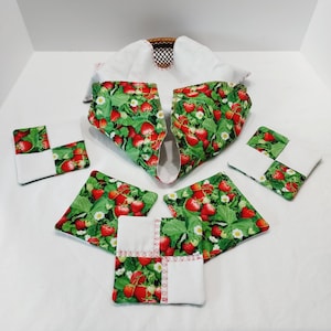 May include: A set of fabric kitchen accessories featuring a strawberry print with red berries, green leaves, and white flowers. The set includes a basket liner with a white interior and four square coasters with white fabric corner accents.